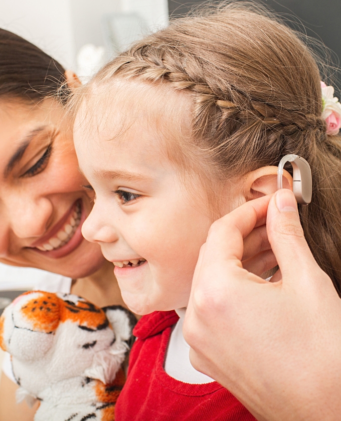 Fonoaudióloga adaptando aparelho auditivo intracanal na orelha de uma menina sorridente com tranças no cabelo, mãe ao lado sorrindo e segurando a criança, urso de pelúcia tigre no colo, ambiente acolhedor e afetivo, fotografia realista de perto, luz natural suave